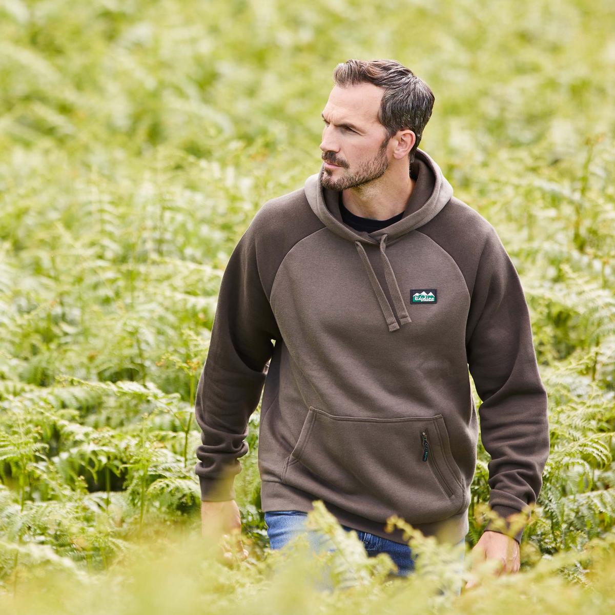 Man wearing a beige and brown hoodie with a kangaroo pocket and a small zippered pocket, featuring a Ridgeline Logo on the upper left side. Walking through a field of plants
