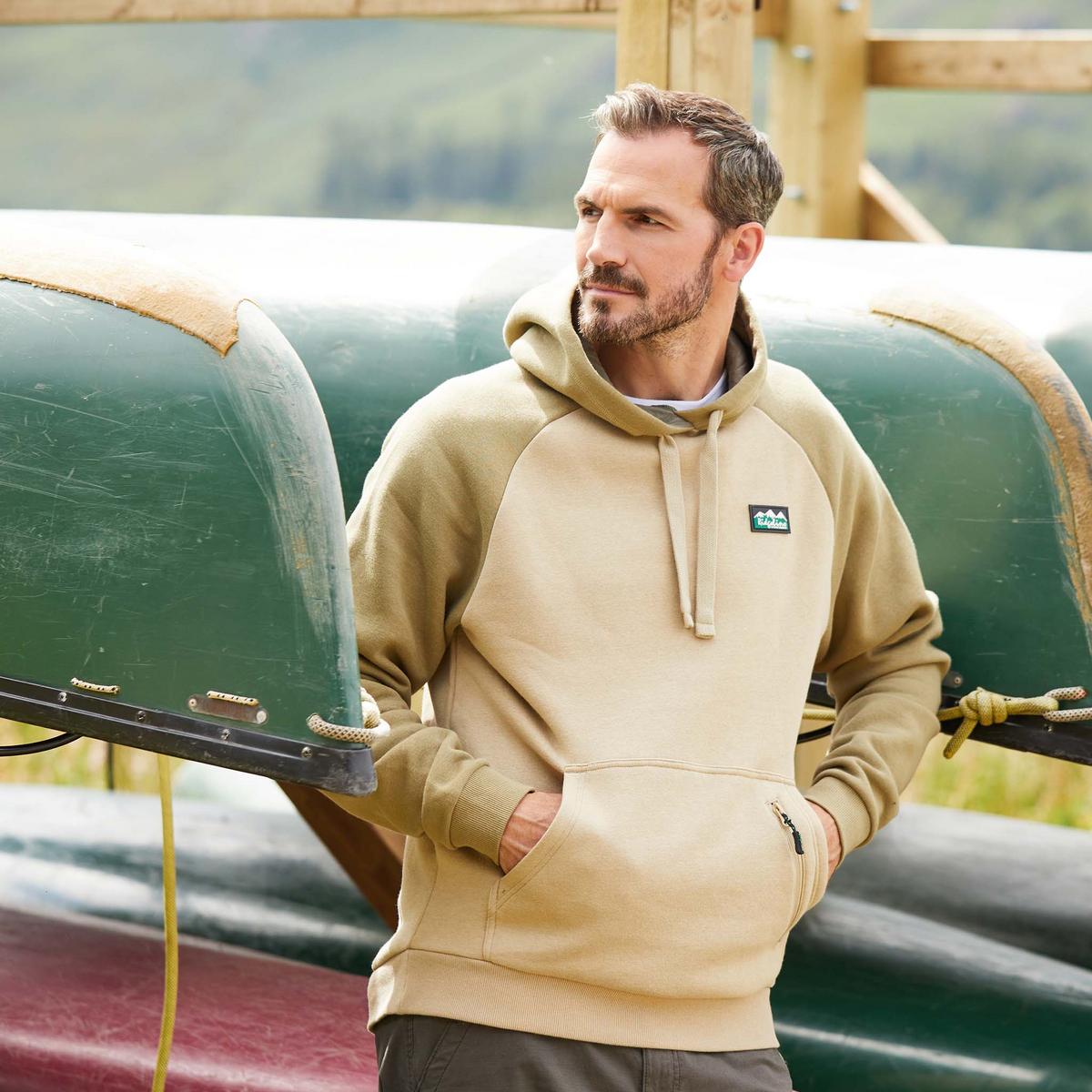 Man wearing a beige and brown hoodie with a kangaroo pocket and a small zippered pocket, featuring a Ridgeline Logo on the upper left side. leaning on Kanoo's