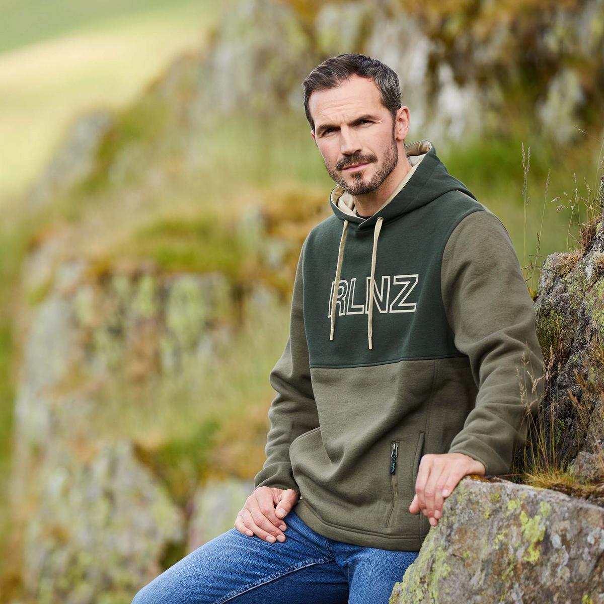 Man wearing a two tone Green hoodie with 'RLNZ' logo outdoors leaning on rocks