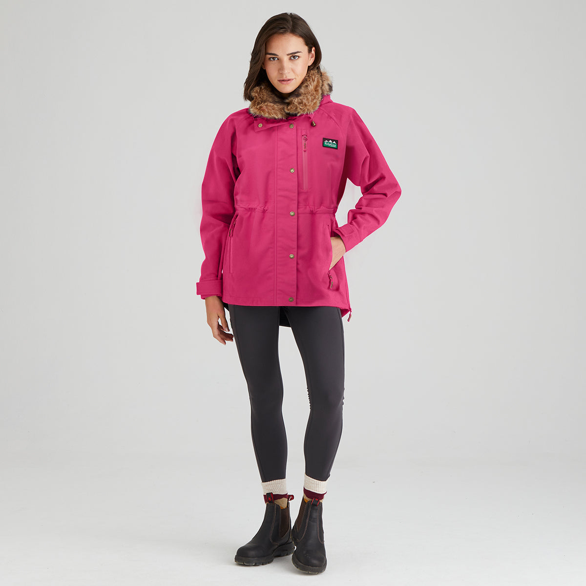 A woman modeling a bright pink outdoor jacket with a brown faux-fur collar, black leggings, and black boots, standing against a light gray studio background.