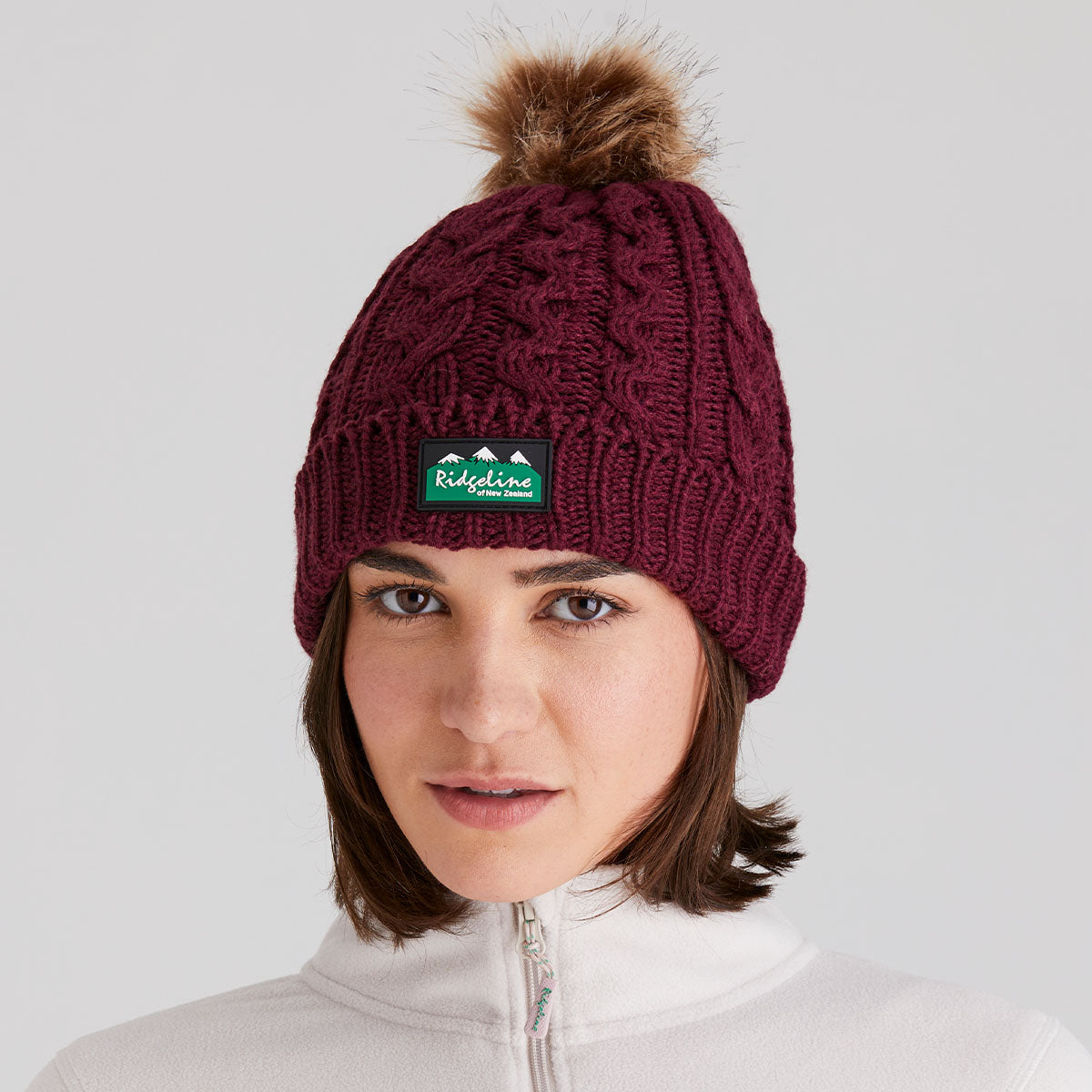 woman wearing a maroon knit beanie with a pom-pom and a visible ridgeline clothing brand logo on a light grey background