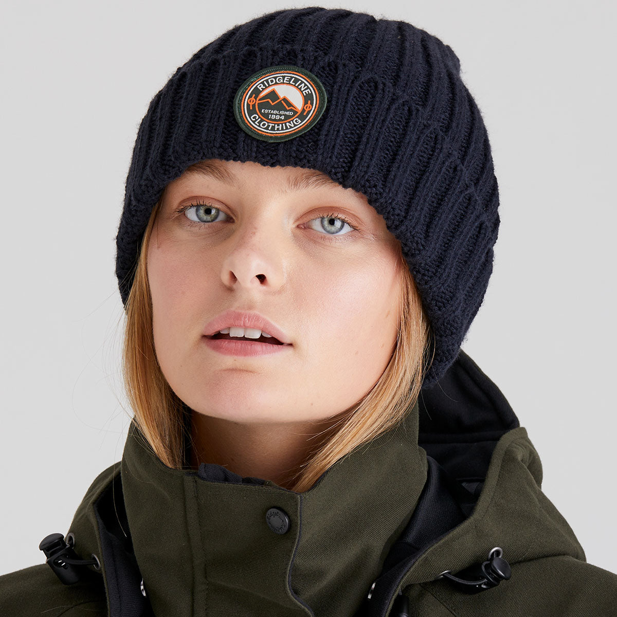 woman wearing a navy knit beanie with a logo and a green coat on a light grey background