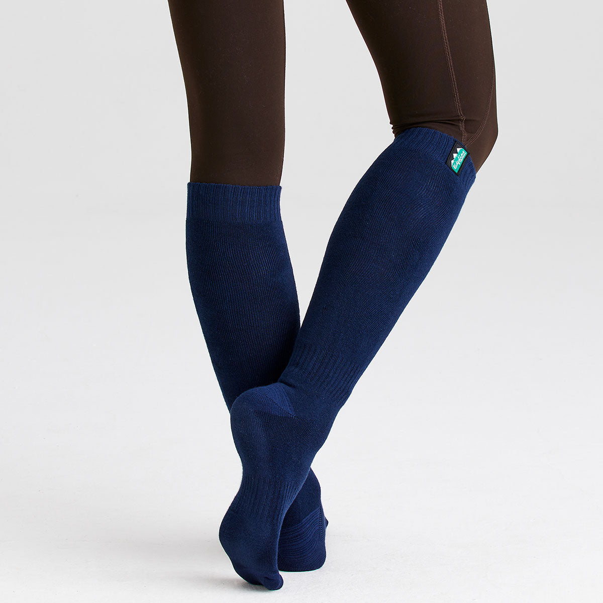 Person wearing knee-high navy blue work socks while standing with crossed legs against a light grey background. The socks feature a ribbed knit design and a small Ridgeline logo tab near the top cuff. The image highlights the fit, length, and appearance of the socks when worn.