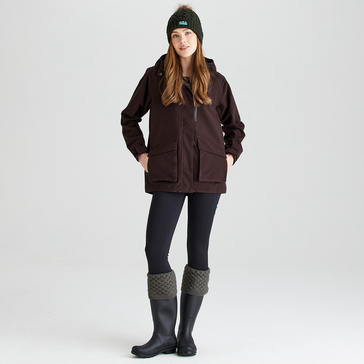 Lifestyle image showing a person wearing olive-green welly socks with chunky cable-knit turnover cuffs styled over dark rubber wellington boots. The outfit is completed with black leggings, a dark outdoor jacket, and a matching olive-green cable-knit bobble hat with a faux-fur pom-pom and Ridgeline logo patch. Photographed against a light grey studio background, the image showcases the coordinated country-inspired look and highlights the welly socks' warmth, comfort, and classic outdoor styling.