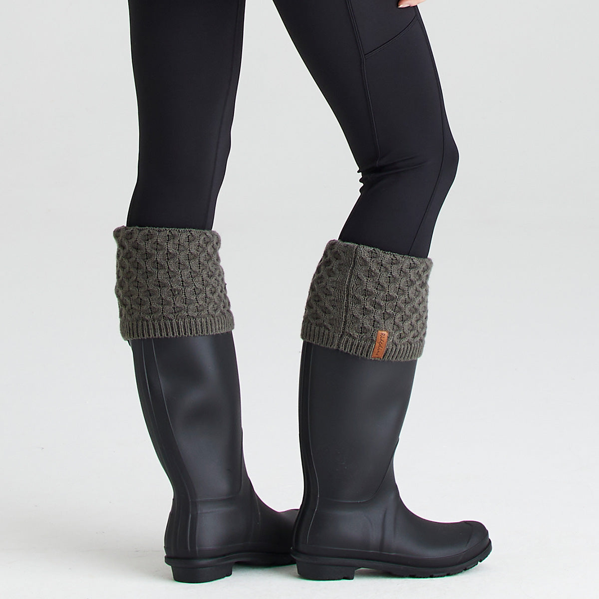 Lifestyle image showing a pair of dark rubber wellington boots worn with olive-green welly sock toppers folded over the boot openings. The welly socks feature a chunky cable-knit pattern and ribbed trim, with a small Ridgeline branded tag visible on one cuff. The boots are paired with black leggings and photographed against a light grey background. The image demonstrates how the knitted welly socks add extra warmth, comfort, and classic country styling when worn with wellington boots.