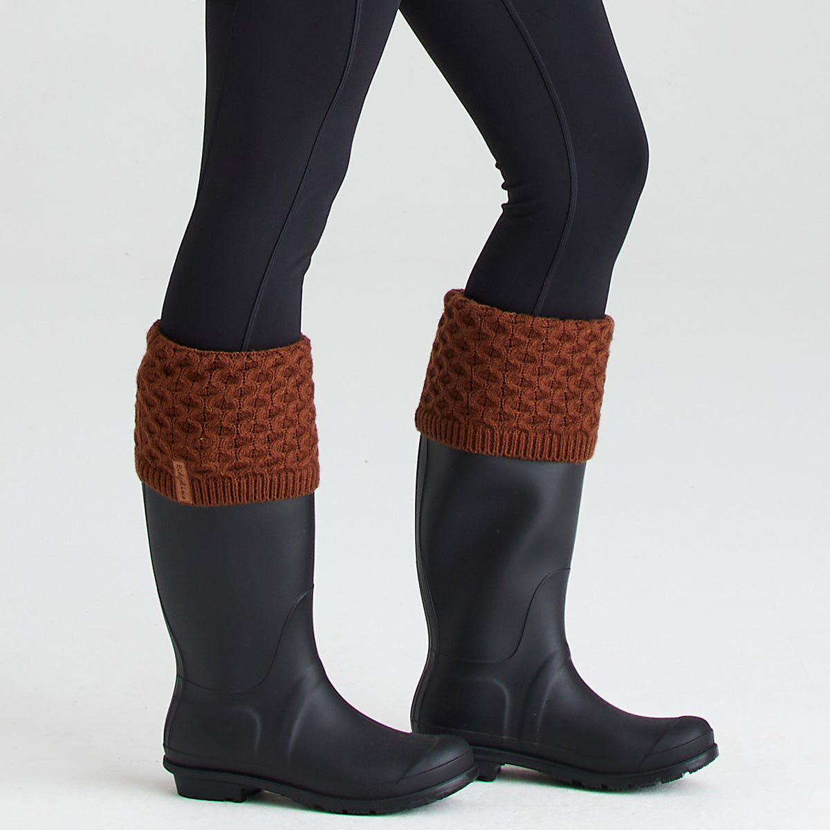 Close-up lifestyle image of a pair of dark rubber wellington boots worn with rust-brown welly sock toppers folded over the boot openings. The welly socks feature chunky cable-knit turnover cuffs with a textured pattern and a small Ridgeline branded tag visible on one cuff. Paired with black leggings and photographed against a light grey studio background, the image highlights the added warmth, comfort, and classic country styling the welly socks provide when worn with wellington boots.