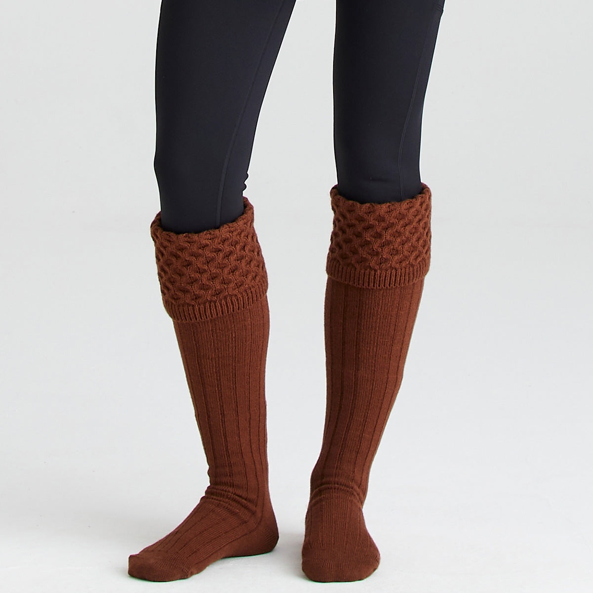 Front view of a pair of rust-brown welly socks with long ribbed legs and chunky cable-knit turnover cuffs, worn with black leggings and displayed against a light grey background. The knee-high socks feature a textured knitted cuff designed to fold over the top of wellington boots, combining classic country styling with added warmth and comfort for cold-weather outdoor wear.