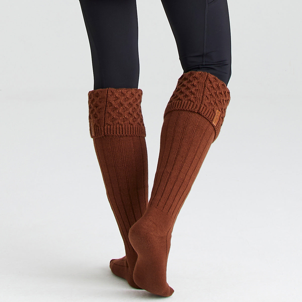 Lifestyle image of a person wearing rust-brown welly socks with long ribbed legs and chunky cable-knit turnover cuffs. The socks are shown without boots and paired with black leggings, highlighting their knee-high length and textured knit construction. A small Ridgeline branded tag is visible on one cuff. Photographed against a light grey studio background, the image showcases the socks' warm, comfortable fit, classic country styling, and suitability for wearing with wellington boots in cold