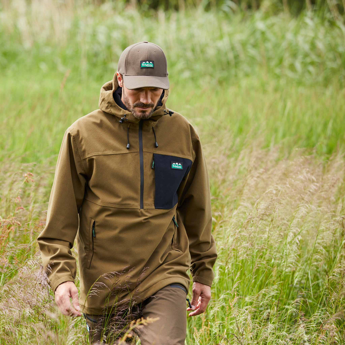 Man wearing a brown waterproof jacket and cap walking through tall grass.