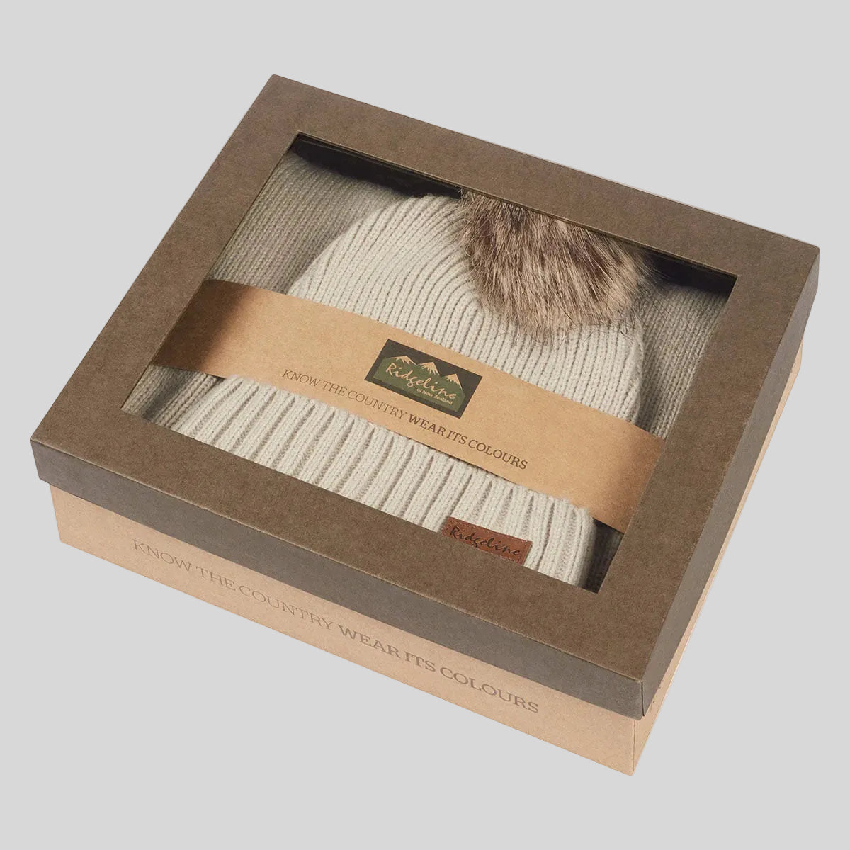 Cream ribbed knit beanie hat with faux fur pom pom in windowed gift box, angled view.