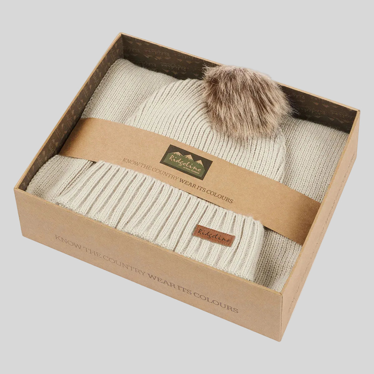 Cream ribbed knit beanie hat with faux fur pom pom in branded gift box, angled view.