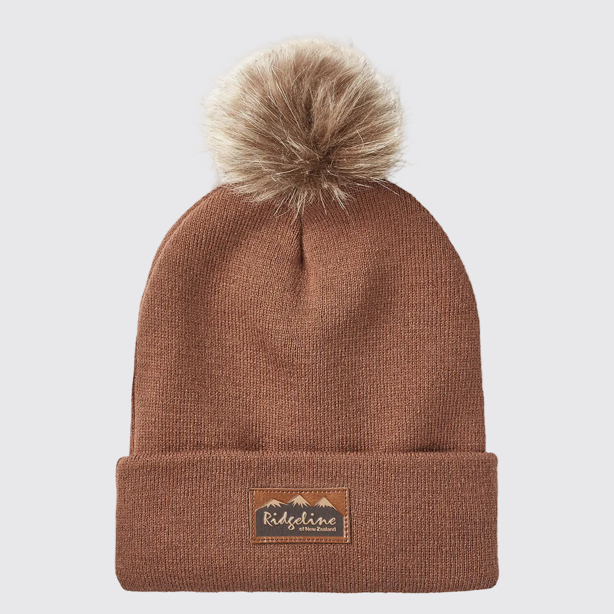 Front view of a rust-brown ribbed knit beanie with a folded cuff and a large faux-fur pom-pom attached at the crown, displayed against a light grey background. A brown faux-leather Ridgeline of New Zealand logo patch featuring a mountain graphic is centered on the cuff. The fine-knit texture, soft pom-pom detail, and stretch-fit construction are clearly visible, highlighting the beanie's warm, comfortable, and stylish cold-weather design.