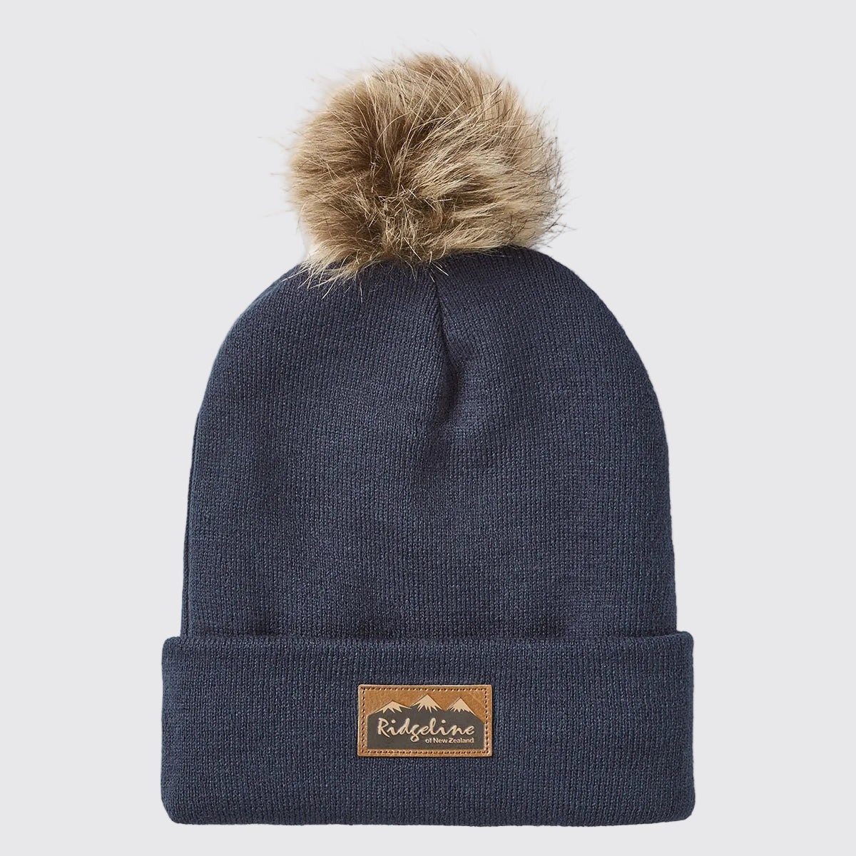 Front view of a navy blue ribbed knit beanie with a folded cuff and a large faux-fur pom-pom attached at the crown, displayed against a light grey background. A brown faux-leather Ridgeline of New Zealand logo patch featuring a mountain graphic is centered on the cuff. The fine-knit texture, soft pom-pom detail, and stretch-fit construction are clearly visible, highlighting the beanie's warm, comfortable, and stylish cold-weather design.