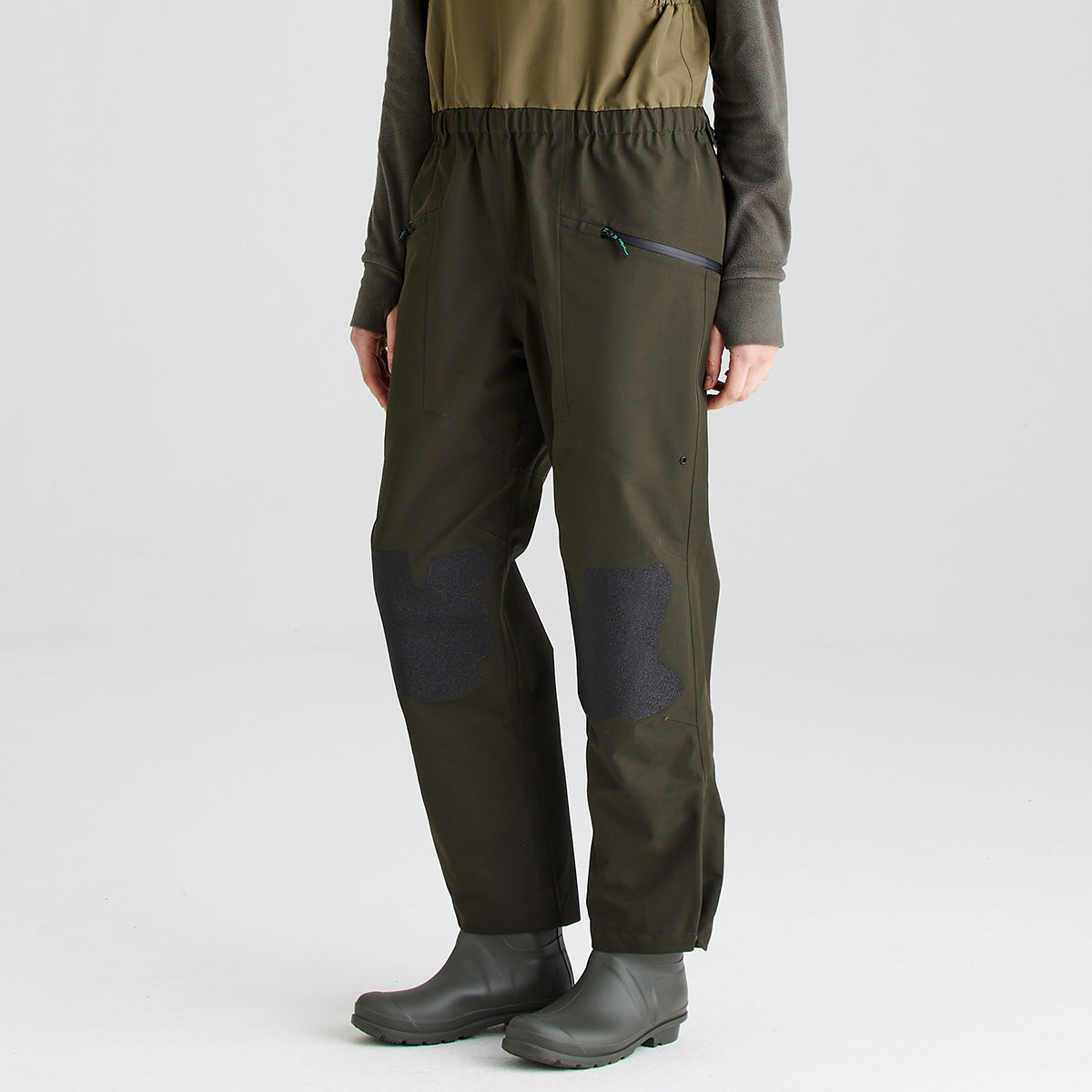 Front view of the lower section of an olive-green waterproof bib and brace with integrated rubber boots, shown against a light gray studio background. The garment features angled zippered pockets at the hips, an elasticated waist, and reinforced knee panels in a contrasting dark textured material for added durability. The image focuses on the leg and knee area, highlighting the practical design and protective construction of the bib and brace.