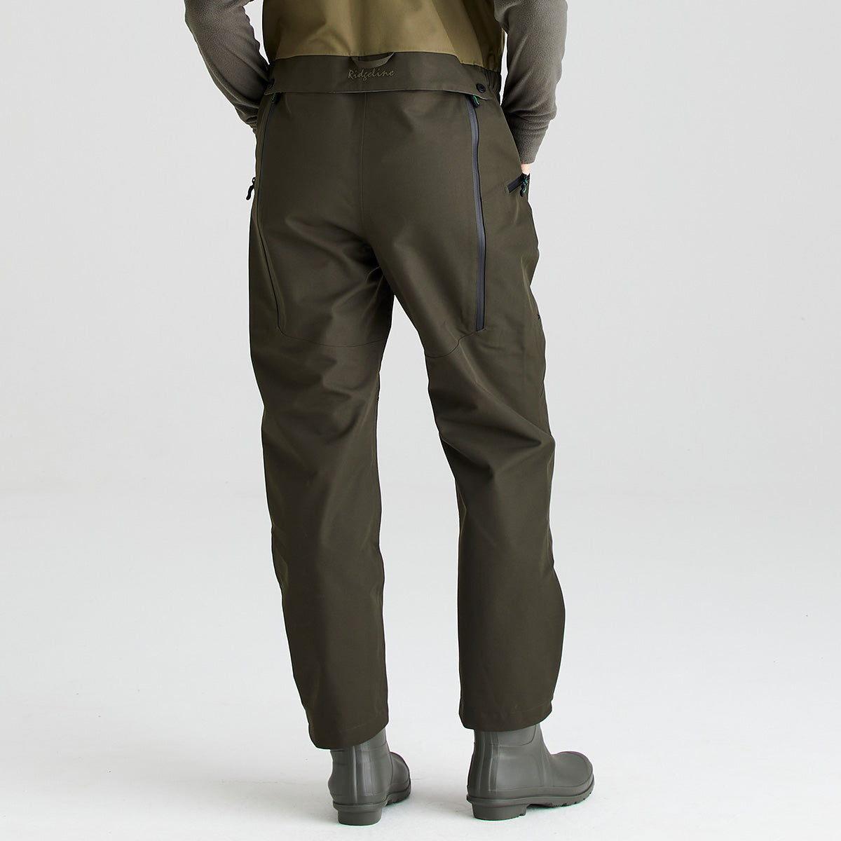 Rear view of an olive-green waterproof bib and brace with integrated rubber boots, shown against a light gray studio background. The garment features a two-tone olive design, adjustable shoulder straps, and a durable waterproof construction. Vertical waterproof zippered pockets are visible on the back, while the straight-leg design and integrated boots provide full lower-body protection. The image highlights the rear fit, pocket placement, and functional outdoor-performance details of the bi