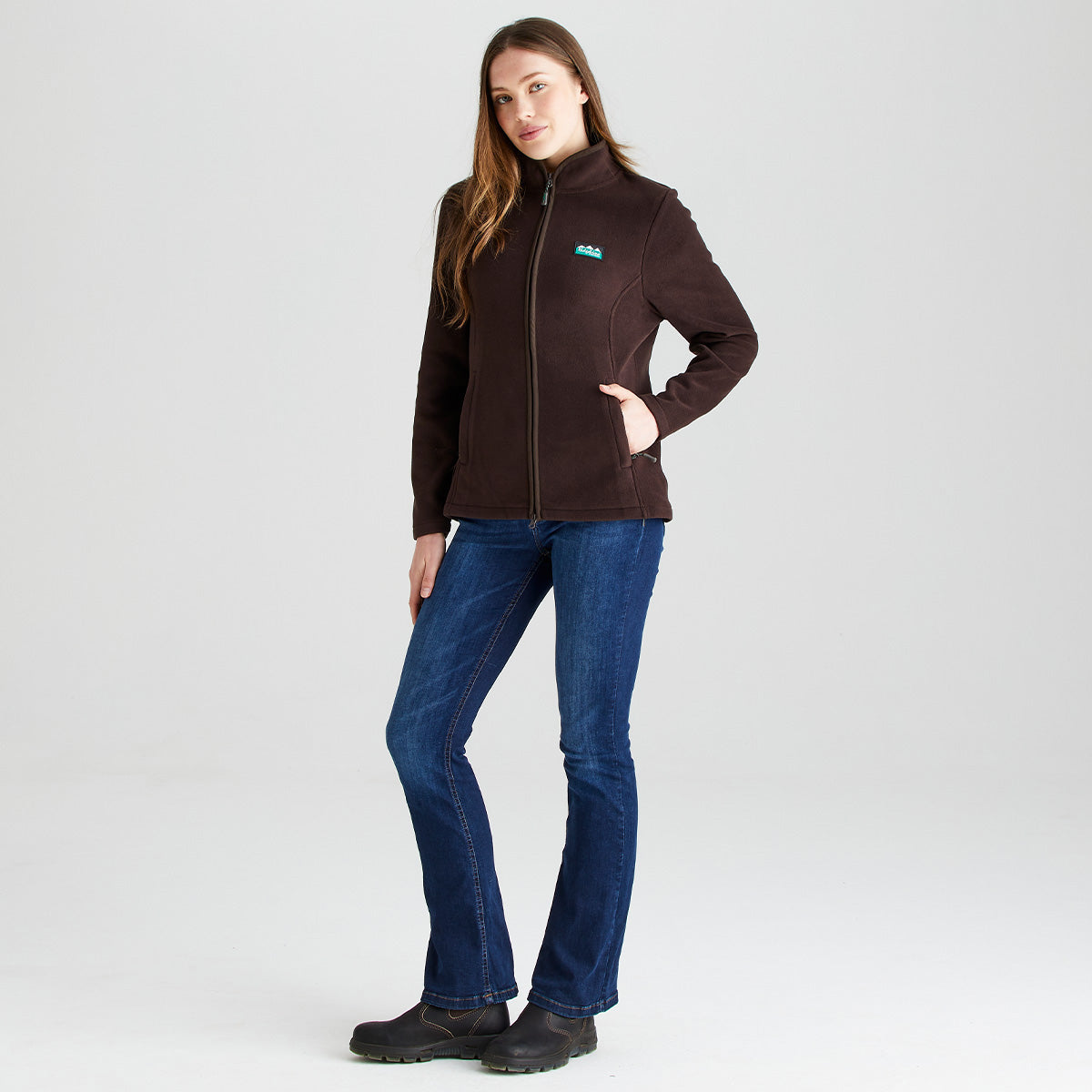 Front view of a dark brown Ridgeline fleece jacket worn with blue jeans. The jacket features a high stand-up collar, full-length front zip fastening, and two zipped hand pockets. A Ridgeline logo patch is displayed on the left chest. The fitted silhouette and soft fleece fabric create a practical, comfortable layer suitable for outdoor and everyday wear. Photographed against a plain light-grey studio background.