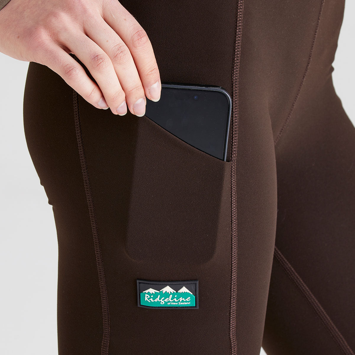 Close-up view of dark brown performance leggings showing a large side thigh pocket. A hand is placing a smartphone into the pocket, demonstrating its storage capacity and easy access. The leggings feature contoured seam detailing and a fitted stretch design, with a branded patch positioned below the pocket. Photographed against a plain light-grey studio background, the image highlights the practical utility pocket and streamlined fit.