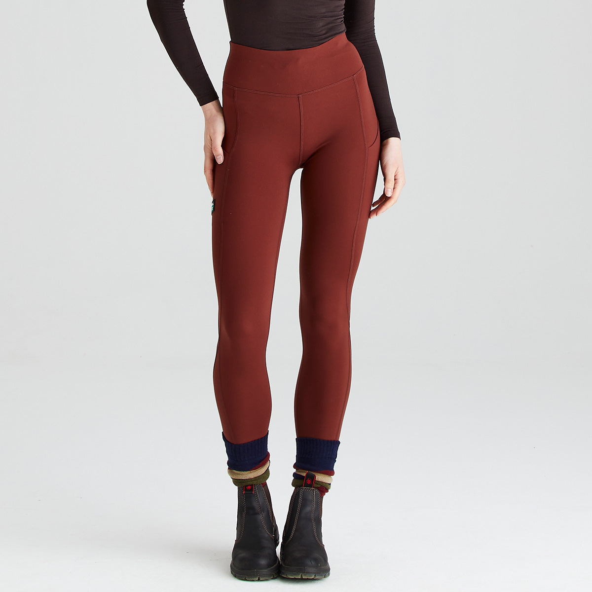 Front view of rust-red high-waisted performance leggings worn with black ankle boots and navy socks with striped cuffs. The leggings feature a wide supportive waistband, streamlined contour seams, and side thigh pockets that blend into the design. The close-fitting stretch fabric creates a sleek silhouette, while the studio image against a plain light-grey background highlights the leggings’ comfort-focused fit and practical activewear styling.