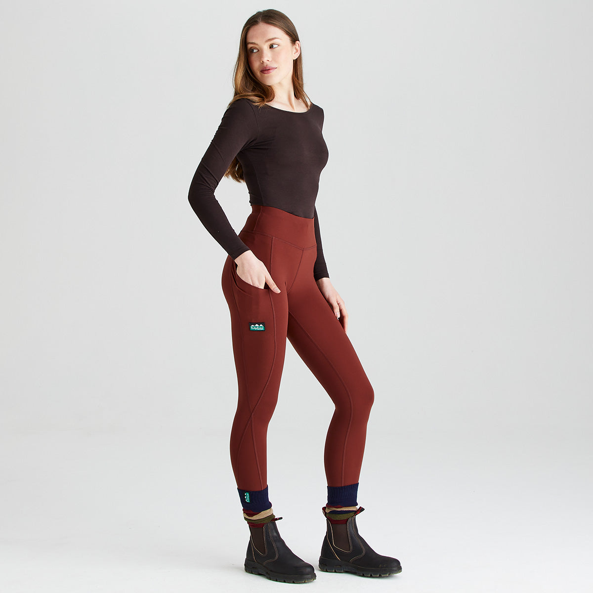 Full-length studio image of a person wearing rust-red high-waisted performance leggings, a fitted dark long-sleeve top, navy socks with striped cuffs, and black ankle boots. The leggings feature a wide supportive waistband, curved contour seams, and a large side thigh pocket, shown with a hand resting inside to demonstrate functionality. A small branded logo patch is visible on the upper thigh. The person is posed in a three-quarter side view against a plain light-grey background, highlighti