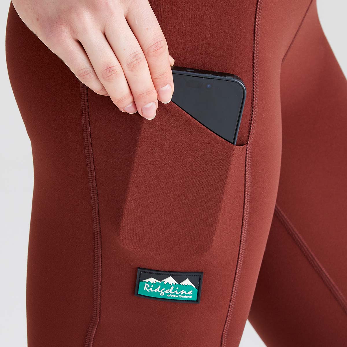 Close-up view of rust-red performance leggings featuring a large side thigh pocket. A hand is placing a smartphone into the pocket, demonstrating its secure storage capacity and easy accessibility. The leggings have a smooth stretch fabric with contoured seam detailing for a streamlined fit, and a branded Ridgeline logo patch positioned below the pocket. Photographed against a plain light-grey studio background, the image highlights the practical utility pocket and functional activewear design.