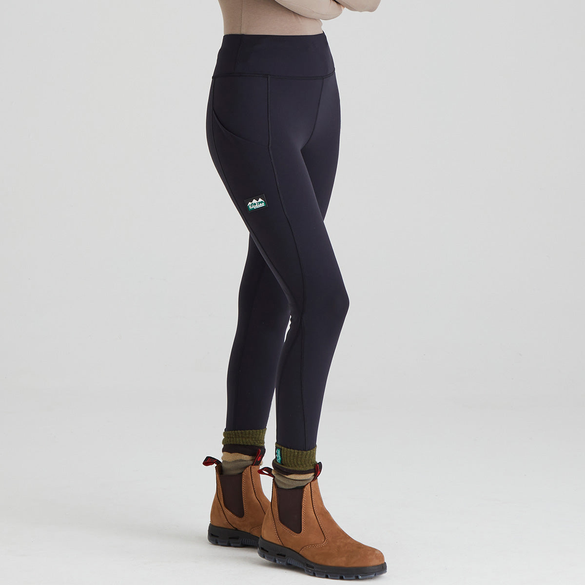woman wearing black leggings with pockets & a ridgeline clothing logo and brown boots on a light grey background