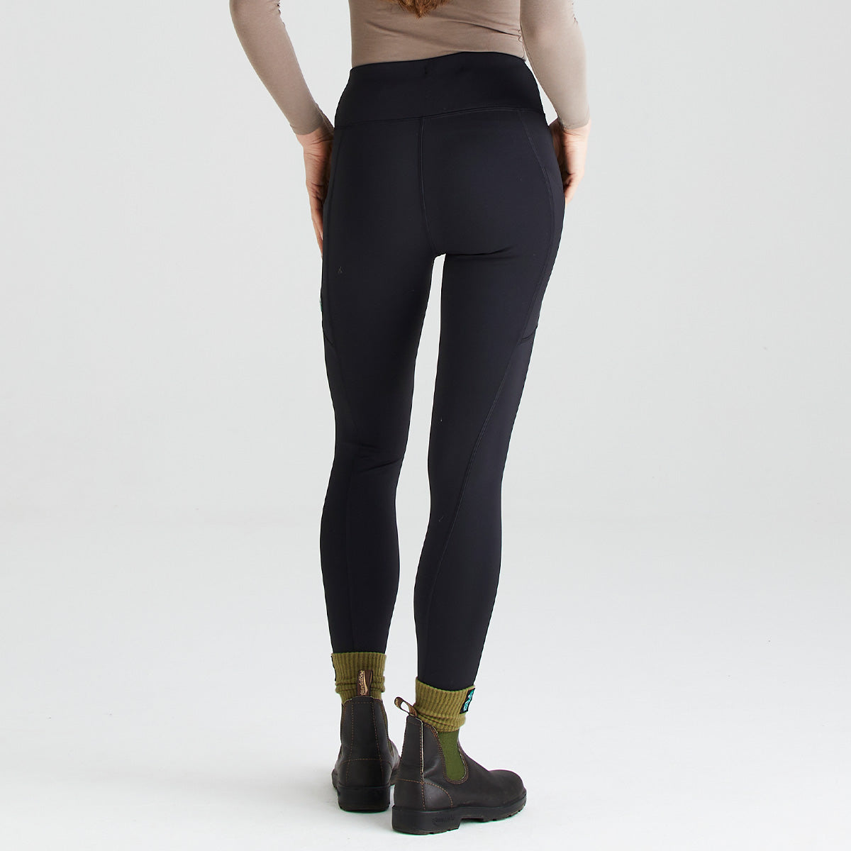 Rear three-quarter view of a person wearing black high-waisted performance leggings with a wide supportive waistband and contouring seam details. The leggings are paired with olive-green socks, dark ankle boots, and a neutral long-sleeve top. Photographed against a plain light grey studio background, the image highlights the back fit, streamlined silhouette, stretch fabric, and ergonomic design of the leggings for active outdoor wear.