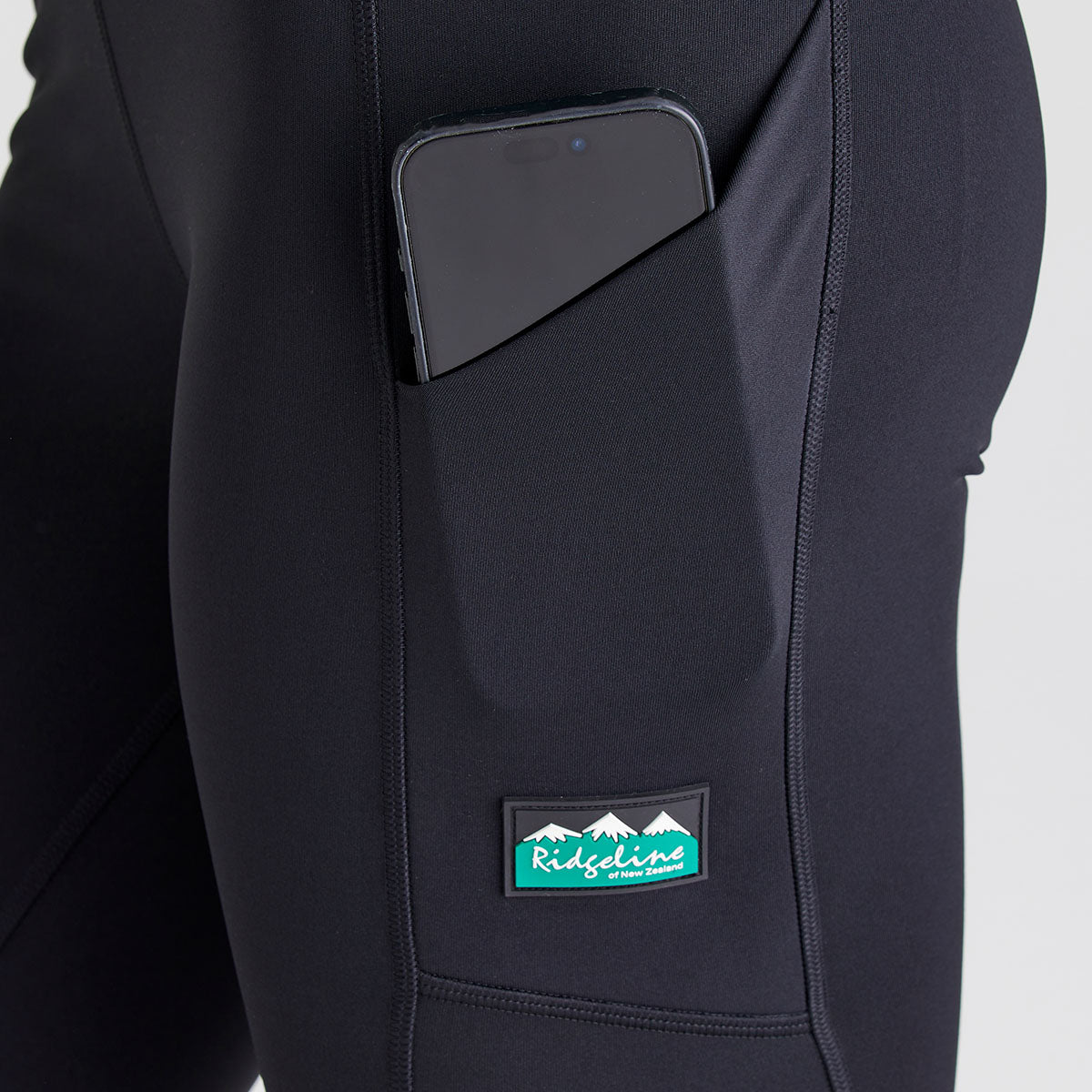 Close-up of black performance leggings showing a side thigh pocket containing a smartphone. The leggings feature smooth stretch fabric, contoured seams, and a fitted design for active outdoor wear. A Ridgeline New Zealand logo patch is stitched below the pocket, highlighting the practical storage feature and branded detailing. Photographed against a plain light grey studio background.