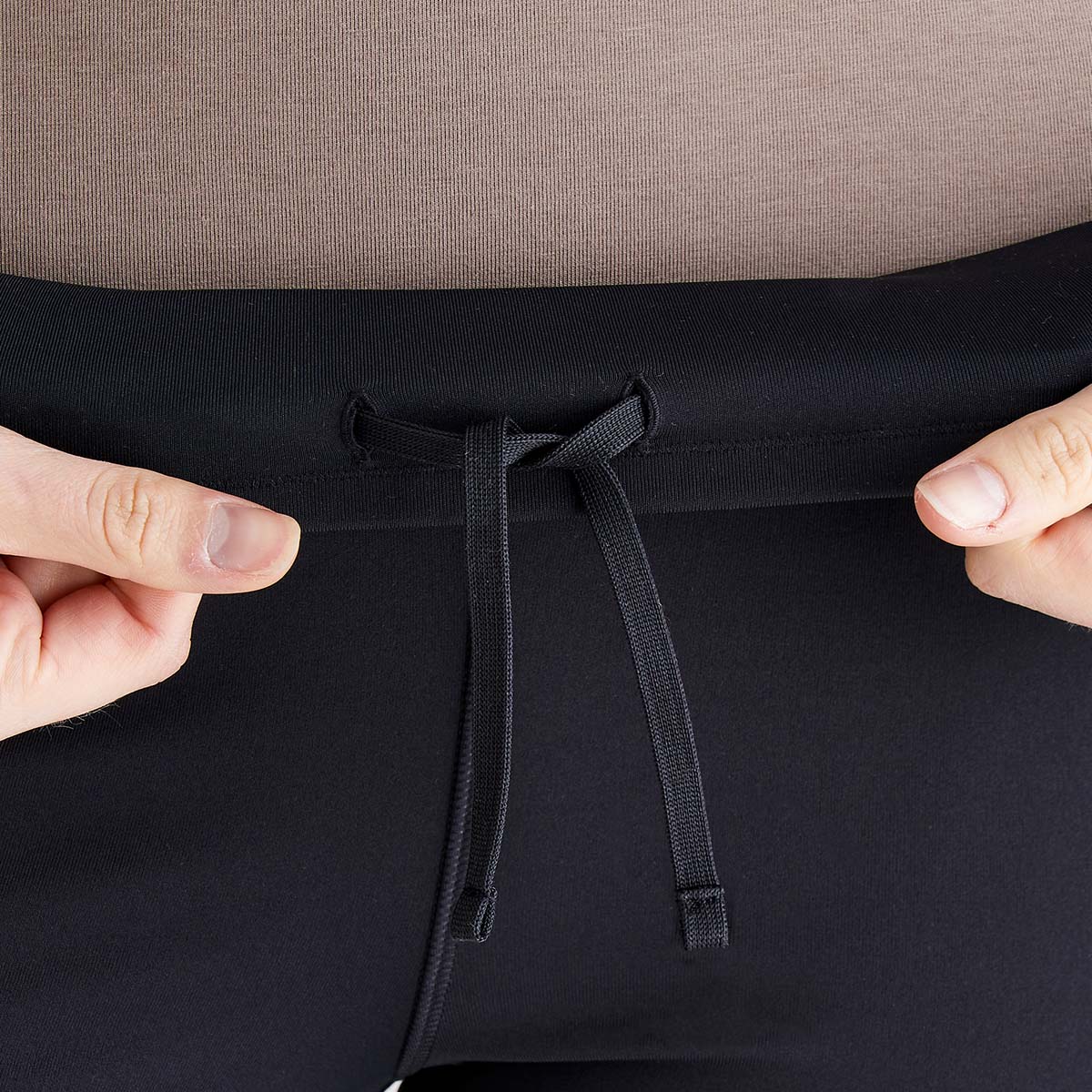 Close-up detail of black performance leggings showing the high-waisted waistband with an internal drawcord tie for a secure, adjustable fit. Two hands are gently stretching the waistband to demonstrate the flexible, supportive stretch fabric. The image highlights the smooth finish, comfort-focused construction, and functional waist design of the leggings.