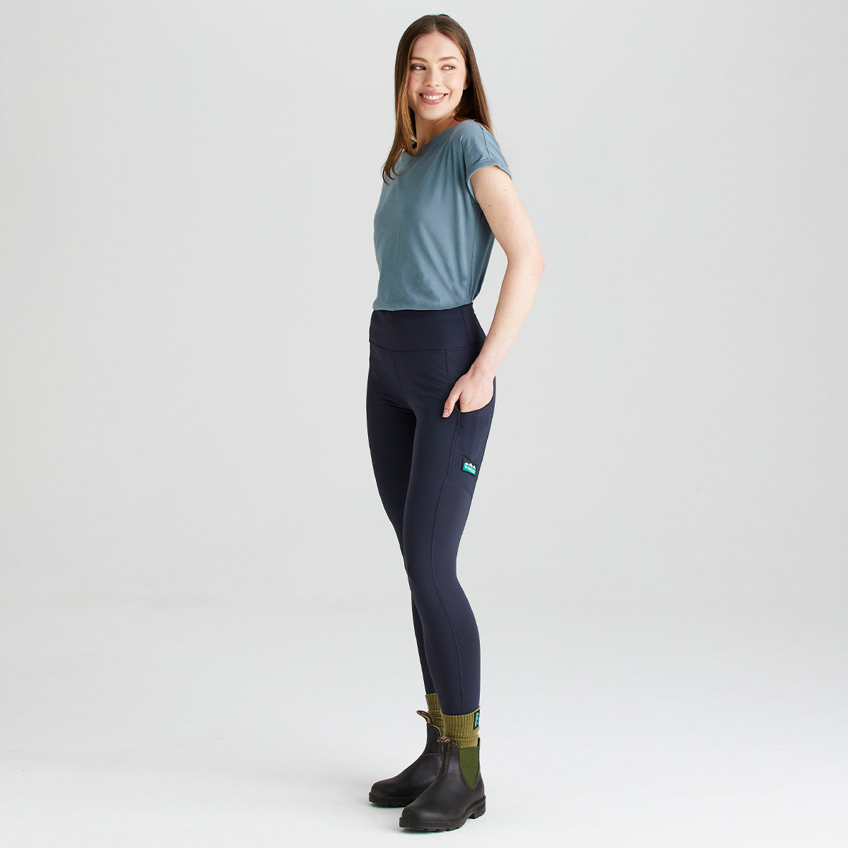 Side-profile studio image of a person wearing Ridgeline navy high-waisted performance leggings with a wide supportive waistband and side thigh pocket. The leggings are paired with a blue short-sleeve top, olive-green socks, and dark ankle boots. A small Ridgeline logo patch is visible on the pocket. Photographed against a plain light grey background, the image highlights the leggings' streamlined fit, stretch fabric, and practical design for outdoor activities and everyday wear.