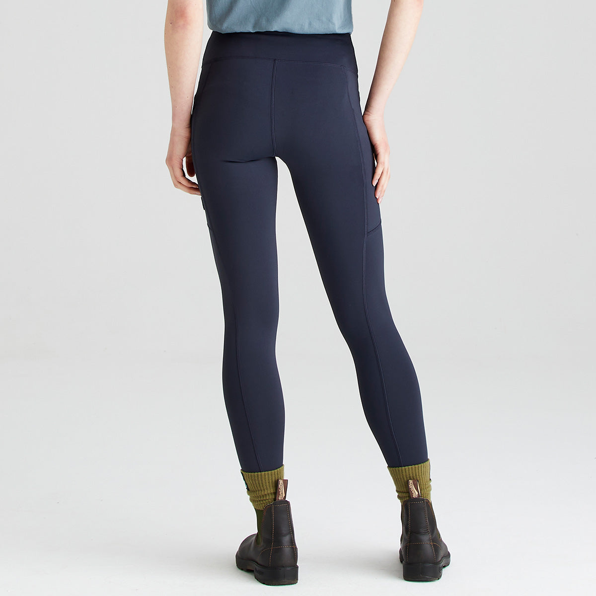 Rear view of a person wearing Ridgeline navy infinity pro high-waisted performance leggings with a wide supportive waistband and side thigh pockets. The leggings are paired with olive-green socks, dark ankle boots, and a blue short-sleeve top. Photographed against a plain light grey studio background, the image highlights the streamlined back fit, stretch fabric, contouring seam details, and practical outdoor-ready design of the leggings.
