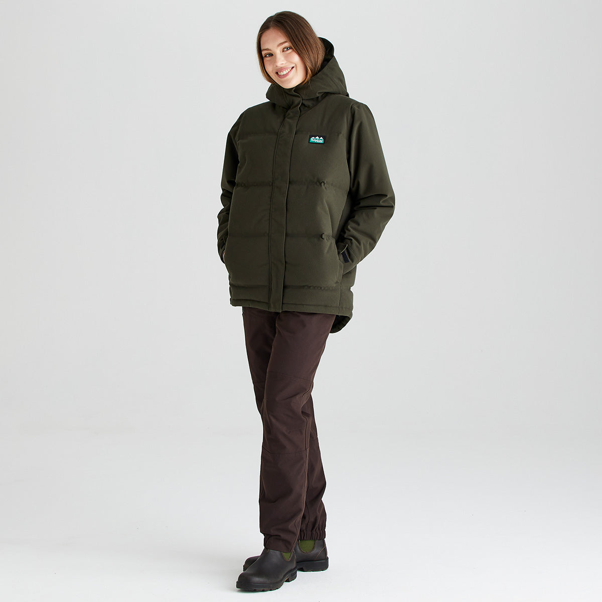 Person wearing a Ridgeline olive green insulated hooded jacket with a full-length front zip, padded construction and side pockets, paired with dark trousers and outdoor boots, shown against a light grey studio background.