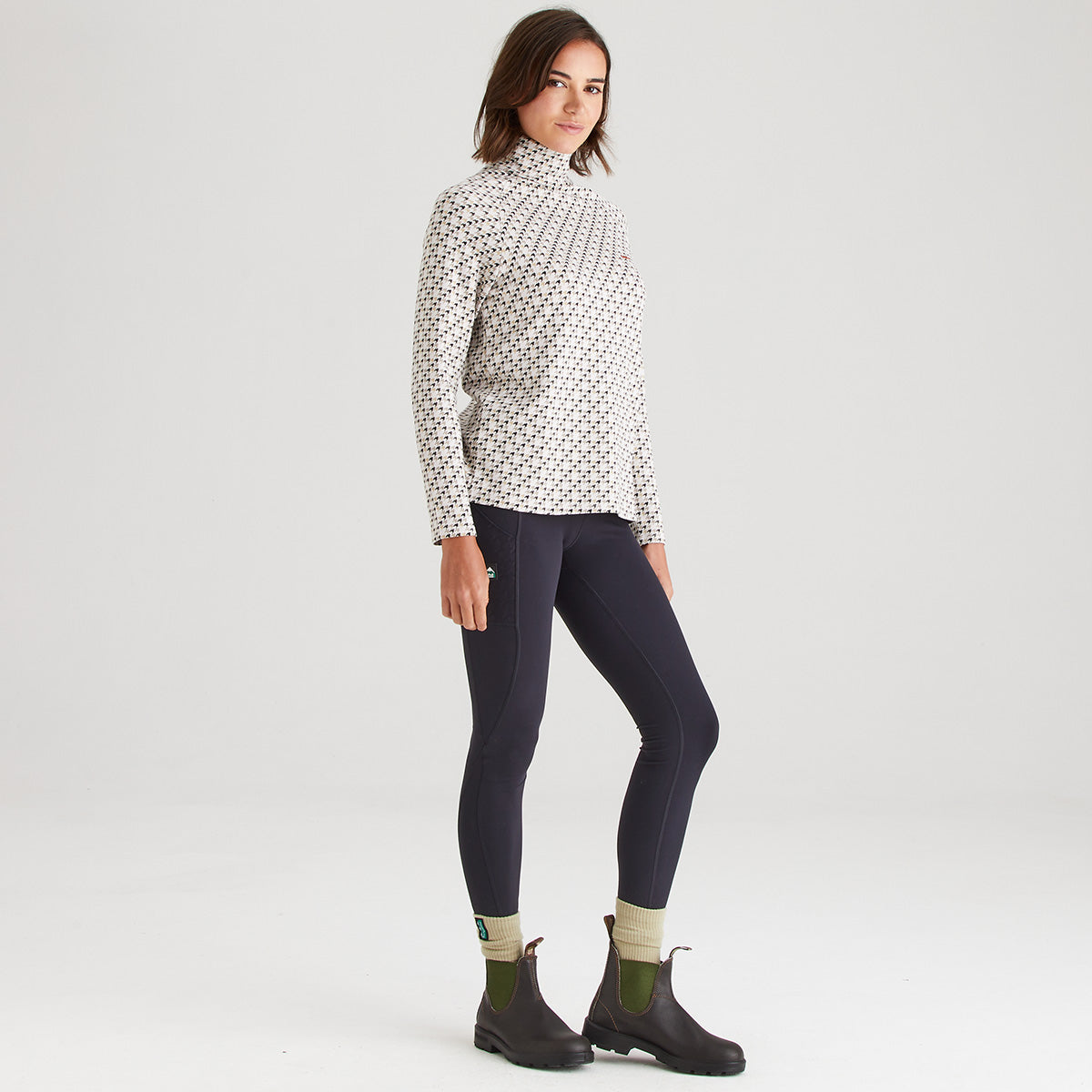 Woman wearing a patterned long-sleeve top, dark leggings, and black boots on a white background