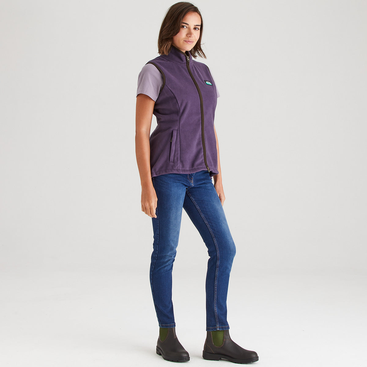 Woman wearing a purple vest over a t-shirt, blue jeans, and black shoes on a white background