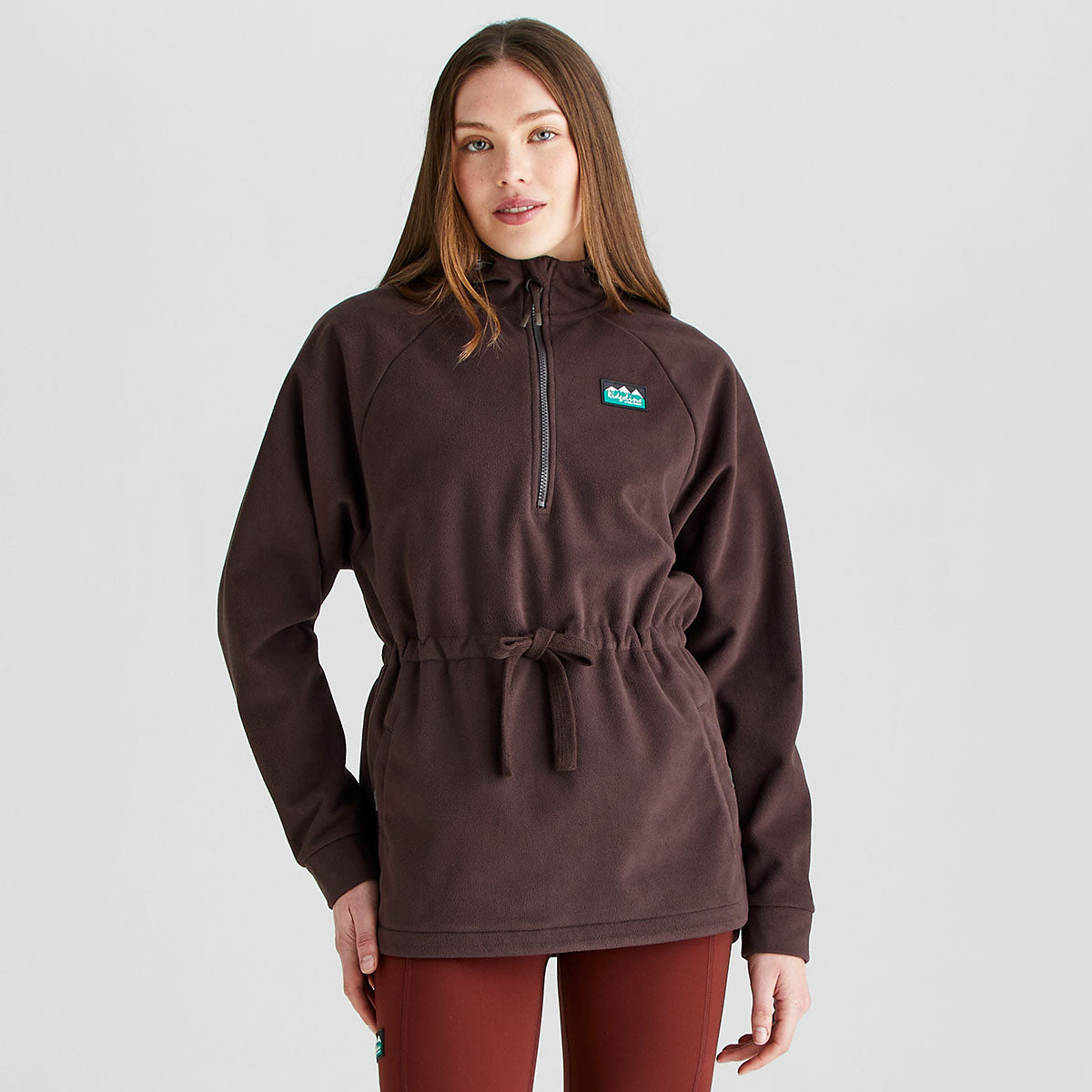 Front view of a person wearing a Ridgeline dark brown or burgundy fleece smock with a half-zip funnel neck, long sleeves, and an adjustable drawcord waist. The fleece features a Ridgeline logo patch on the chest and a soft brushed finish for warmth and comfort. Paired with rust-coloured leggings, the garment is photographed against a plain light grey studio background, highlighting the relaxed fit and practical outdoor styling.