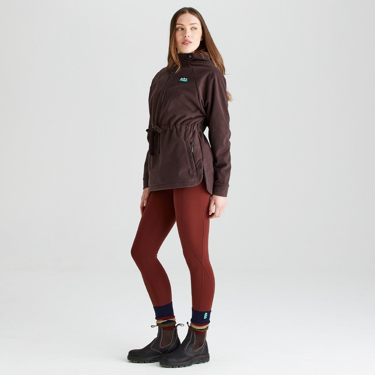 Three-quarter full-length studio image of a person wearing a Ridgeline dark brown or burgundy fleece smock with a half-zip funnel neck, adjustable drawcord waist, and zipped hand pockets. The fleece is paired with rust-coloured performance leggings, striped socks, and dark outdoor ankle boots. Photographed against a plain light grey background, the image highlights the smock's relaxed fit, curved hem, soft fleece fabric, and practical outdoor styling.