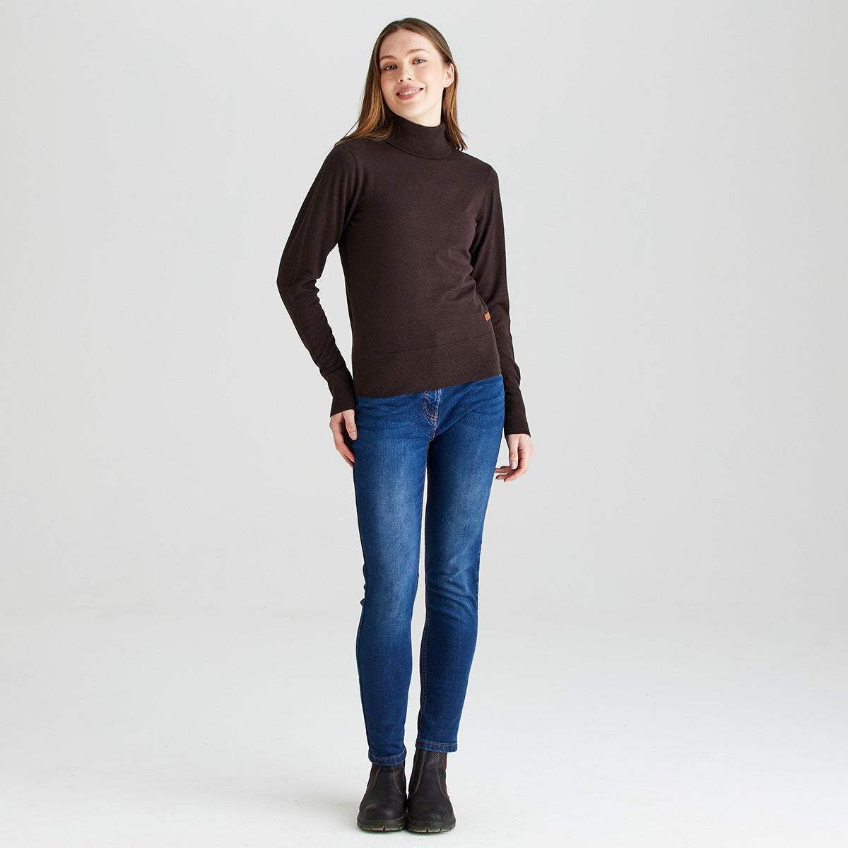 Full-length view of a model wearing a Ridgeline dark brown knitted roll neck jumper with a high turtleneck, long sleeves, and ribbed hem and cuffs. The jumper is paired with blue slim-fit jeans and black ankle boots against a light grey studio background.
