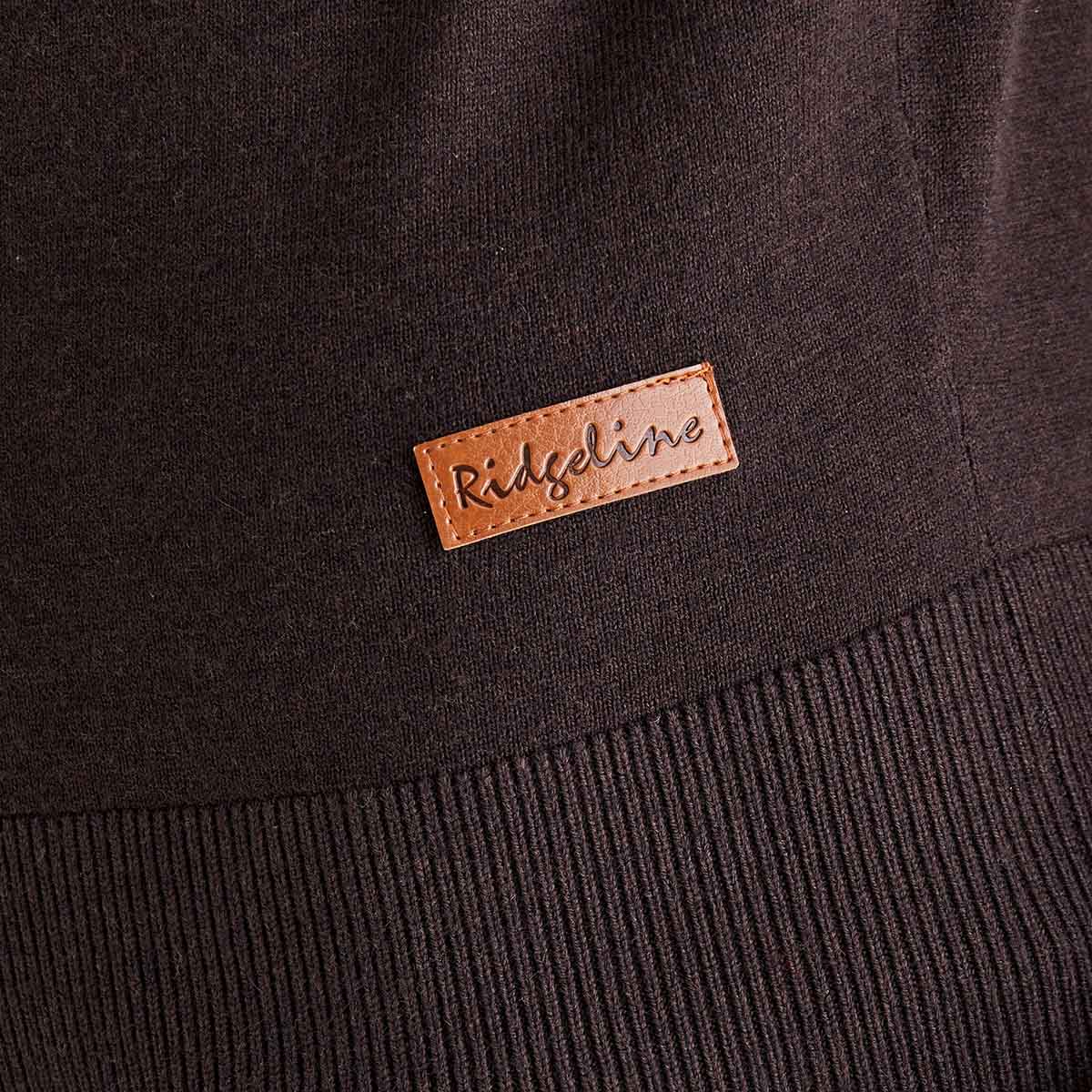 Close-up detail of a dark brown Ridgeline knitted roll neck jumper, showing the ribbed hem and a stitched tan leather Ridgeline logo patch near the lower front. The fine knit texture and quality finishing are clearly visible.