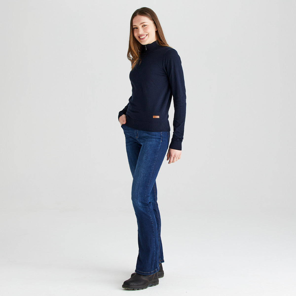 Full-length studio image of a person wearing a navy blue quarter-zip knit sweater, blue jeans, and dark ankle boots. The sweater features a high collar, fine-knit construction, ribbed hem and cuffs, and a small brown Ridgeline logo patch near the hem. The person is shown in a side-profile pose against a plain light gray background, highlighting the fit, silhouette, and styling of the garment.