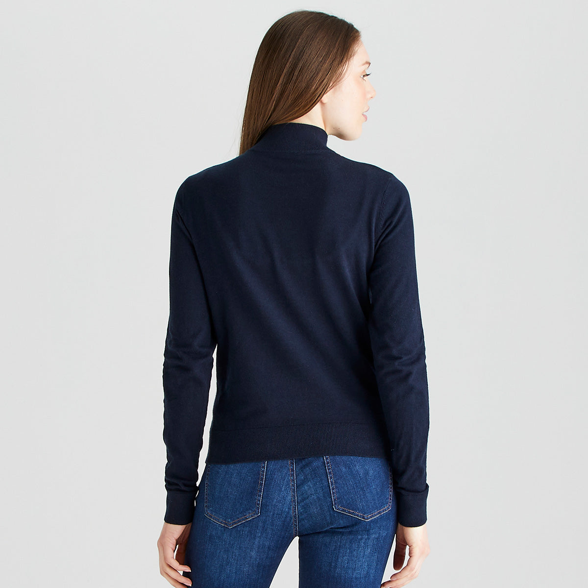 Rear view of a person wearing a navy blue quarter-zip knit sweater and blue jeans against a plain light gray studio background. The sweater features a high collar, fine-knit construction, long sleeves, and a ribbed hem. The image highlights the back fit, shape, and clean design of the garment, with straight hair falling down the back and partially covering the upper section of the sweater.