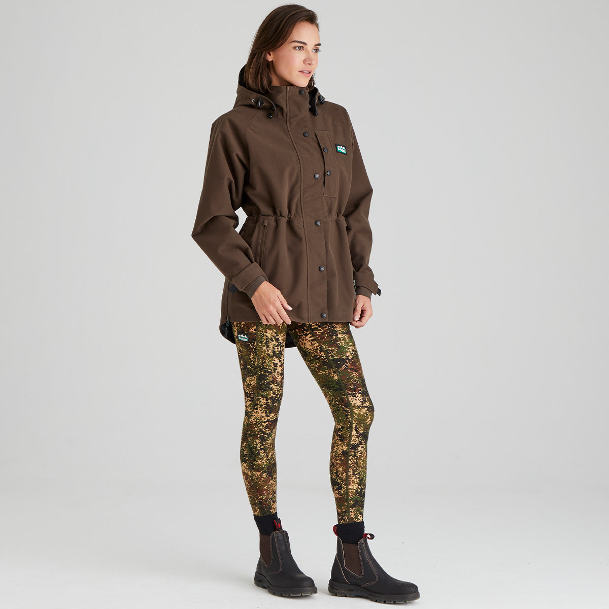 Woman wearing a brown waterproof jacket and camouflage leggings on a grey background