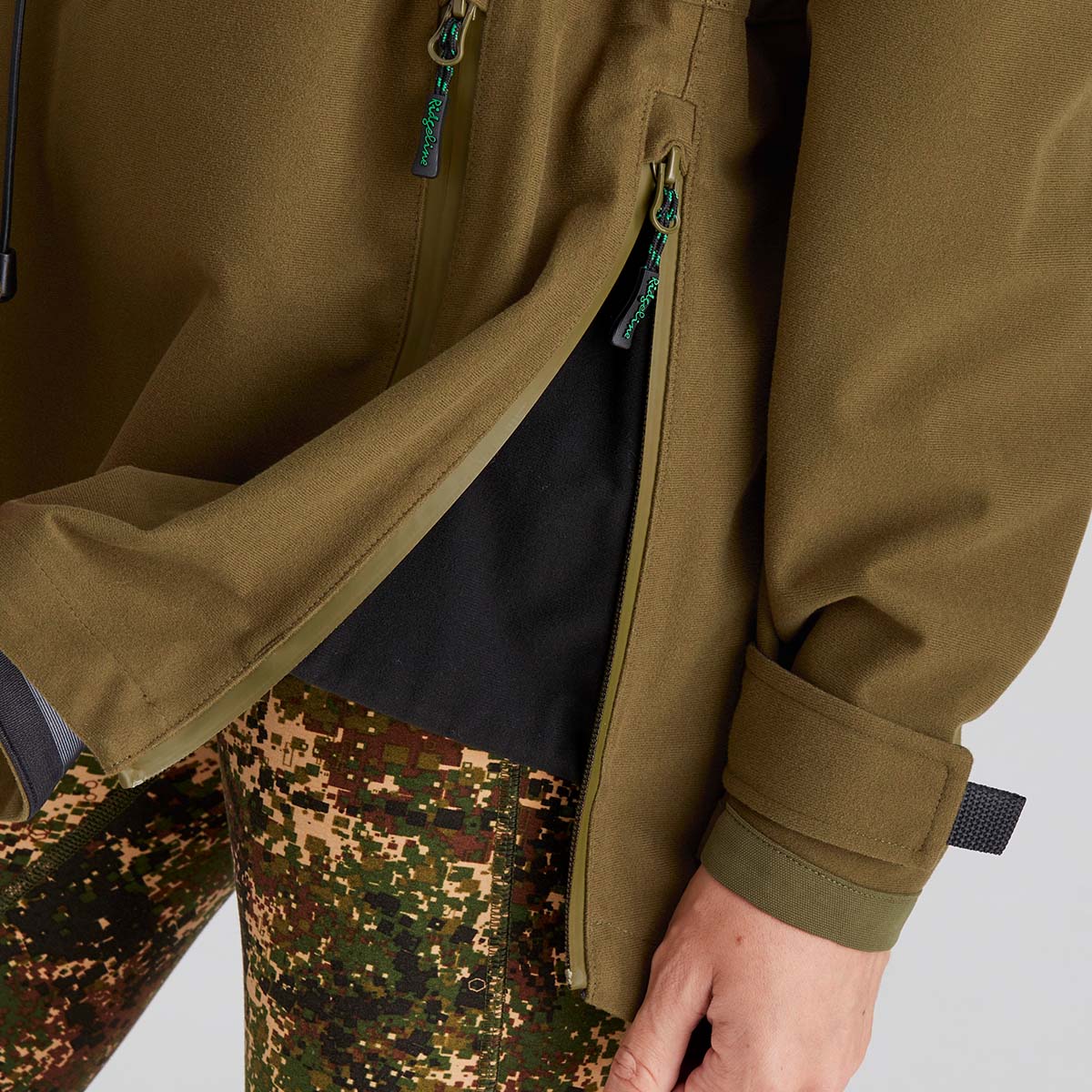 Close-up of a person wearing a brown jacket showing the side zip and camouflage pants on a light grey background