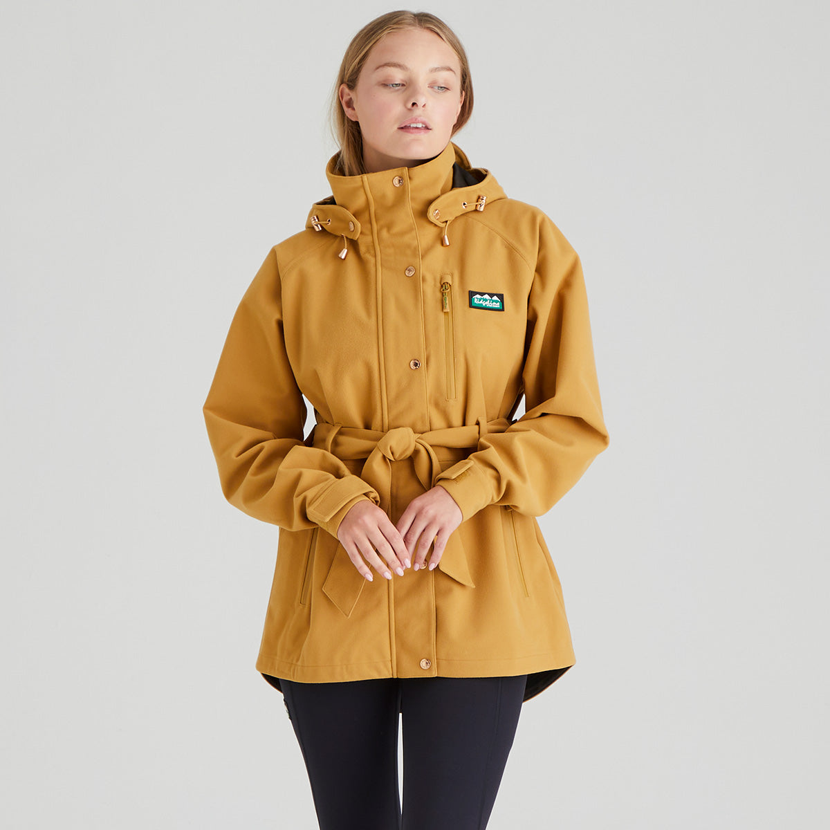 Woman wearing a mustard yellow jacket with a brand logo on a grey background