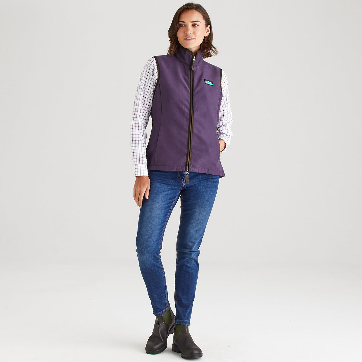 Woman wearing a purple vest over a checkered shirt with blue jeans on a white background