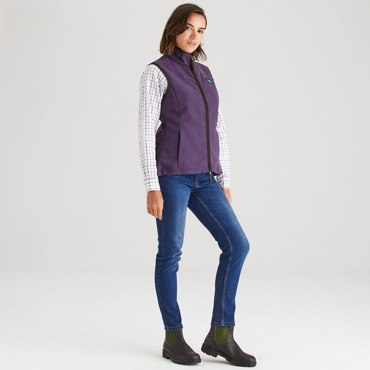 Woman wearing a purple vest over a checkered shirt, blue jeans, and black boots on a white background