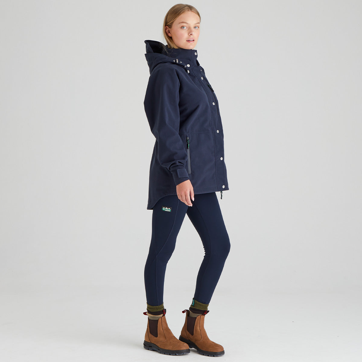 A woman modeling a navy blue hooded jacket, navy leggings, and brown Chelsea boots against a light gray studio background.