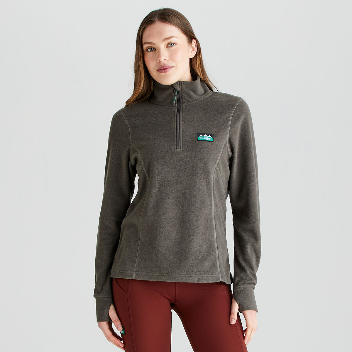 Front view of a grey Ridgeline fleece pullover worn with rust-red leggings. The pullover features a high stand-up collar, a quarter-zip neckline, long sleeves, and a soft fleece construction for lightweight warmth and comfort. A Ridgeline logo patch is positioned on the left chest, while shaped seam detailing creates a streamlined fit. Photographed against a plain light-grey studio background, the image highlights the pullover’s practical outdoor style and layering versatility.