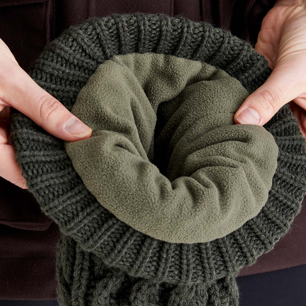 Close-up image of the inside of a dark charcoal cable-knit beanie being held open with both hands to display the interior lining. The beanie features a soft olive-green fleece lining that covers the inside for added warmth and comfort. The ribbed cuff and chunky cable-knit outer texture are clearly visible around the opening, highlighting the hat's insulated construction and cold-weather design. The image focuses on the contrast between the textured knit exterior and the plush fleece interio