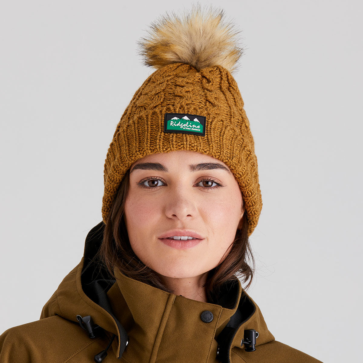 Person wearing a brown knit beanie with a pom-pom and a visible Ridgeline Clothing brand logo, against a plain background.