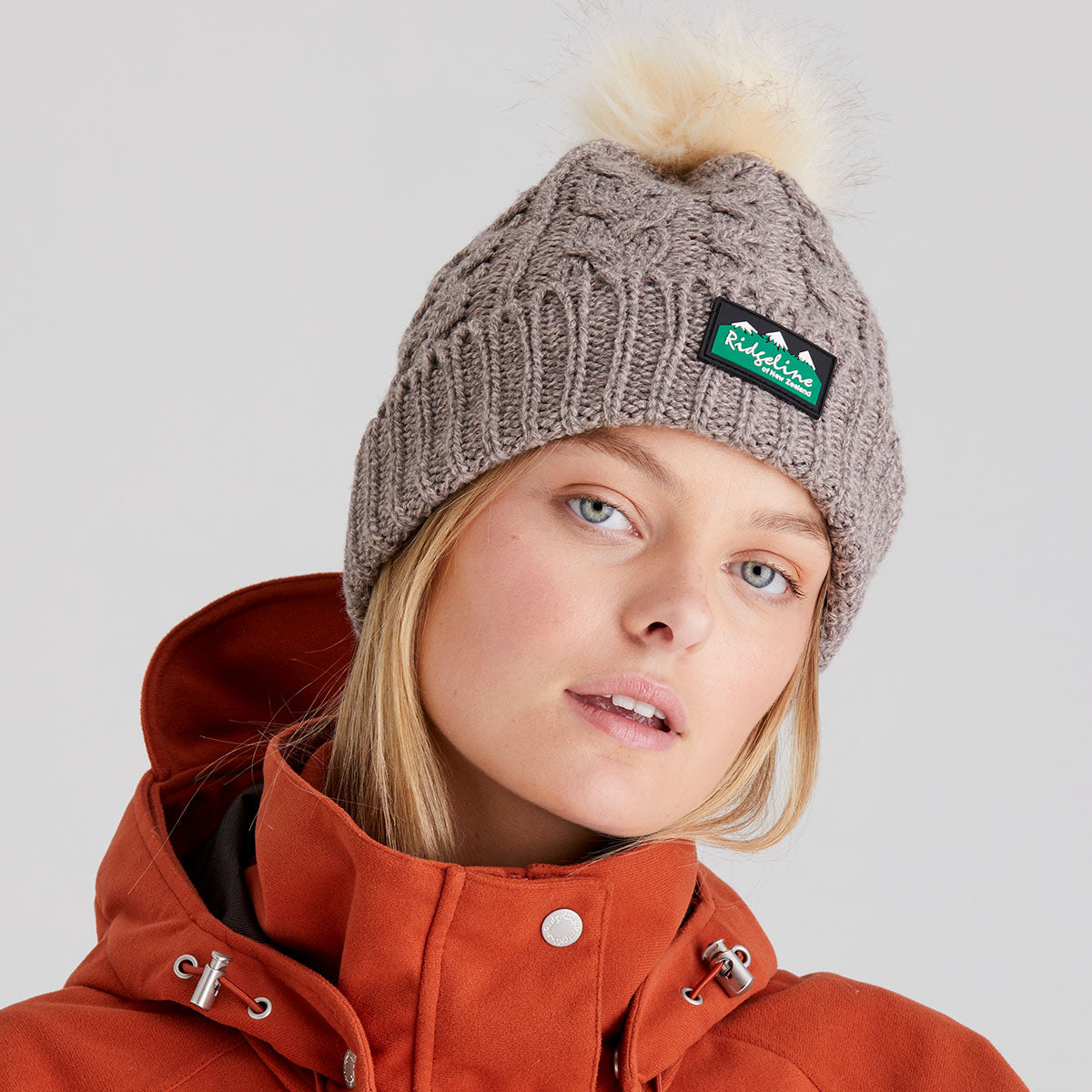 Person wearing a knit beanie with a ridgeline clothing brand logo and an orange jacket on a light grey background