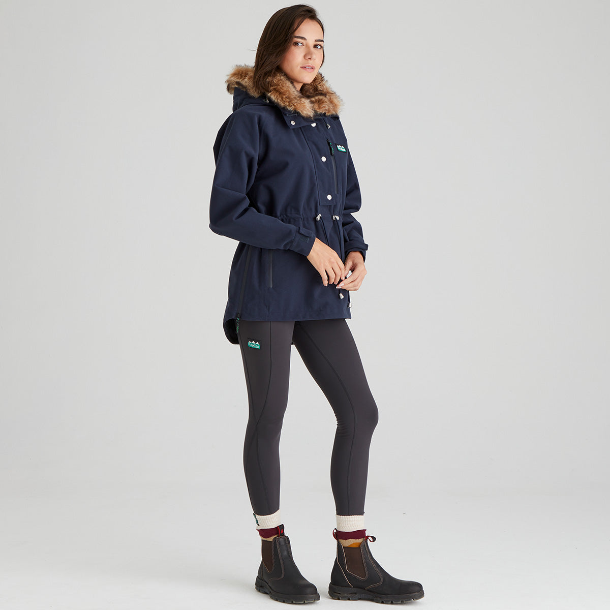 Woman wearing a navy blue jacket with fur hood and dark legging on a light grey background